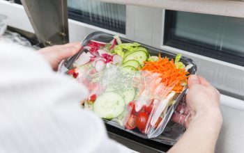 Production line of pre-packaged convenience food. Over the shoulder view, a woman wrapping ready to eat salad in a wrapping machine with cling foil. This image is part of a series.
